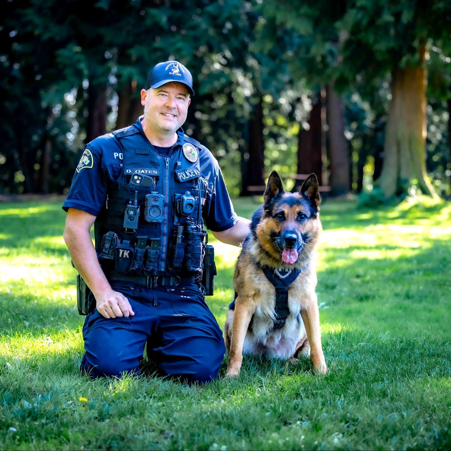 K9 Copper is sitting on the grass next to Officer Derek Oates who is kneeling down and smiling