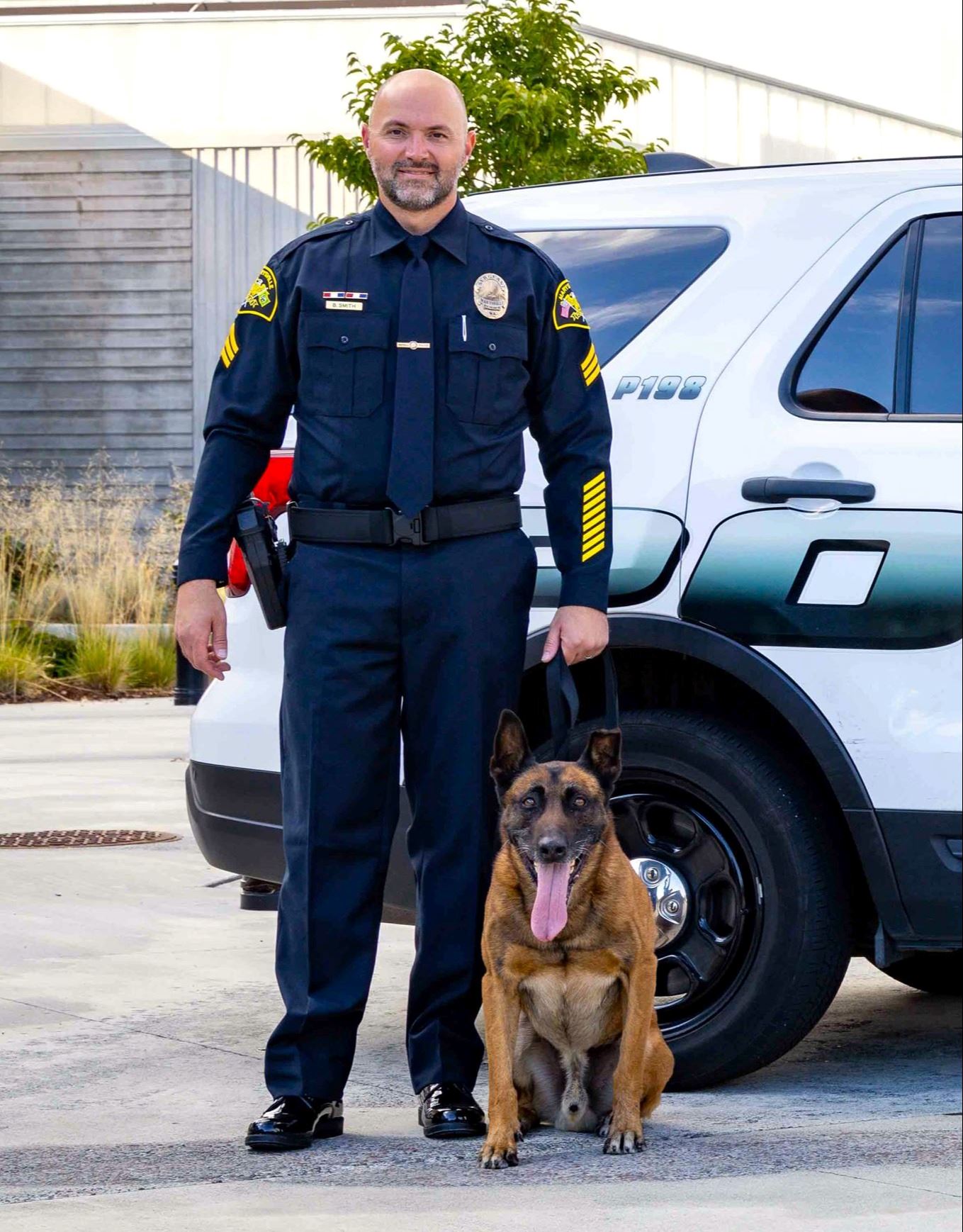 K9 Steele seen sitting in front of patrol car, alongside Officer Brad Smith who is smiling